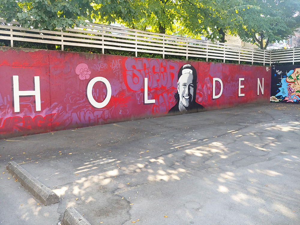 A bold mural across a low, concrete wall features a red and burgundy painted background with “Holden” in white san serif script across the wall. The middle of the mural features a painting of Holden in grey, white and black. He is wearing a backwards ball cap, he has a piercing in one ear and he is smiling. 