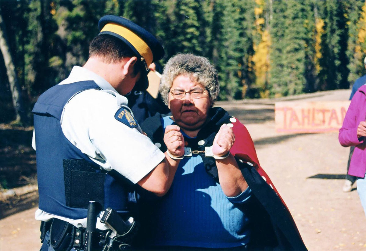 An RCMP officer handcuffs an Elder Indigenous woman wearing a red ceremonial blanket.