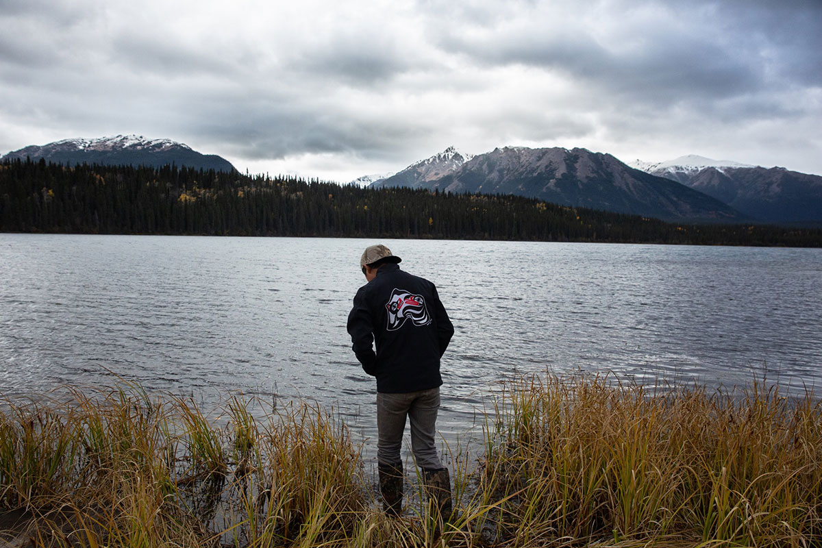 A young teenager is wearing a black jacket with Indigenous insignia on the back. He is wearing rubber boots and stepping near the sacred headwaters. The sky and water is grey. In the background is a mountain range.