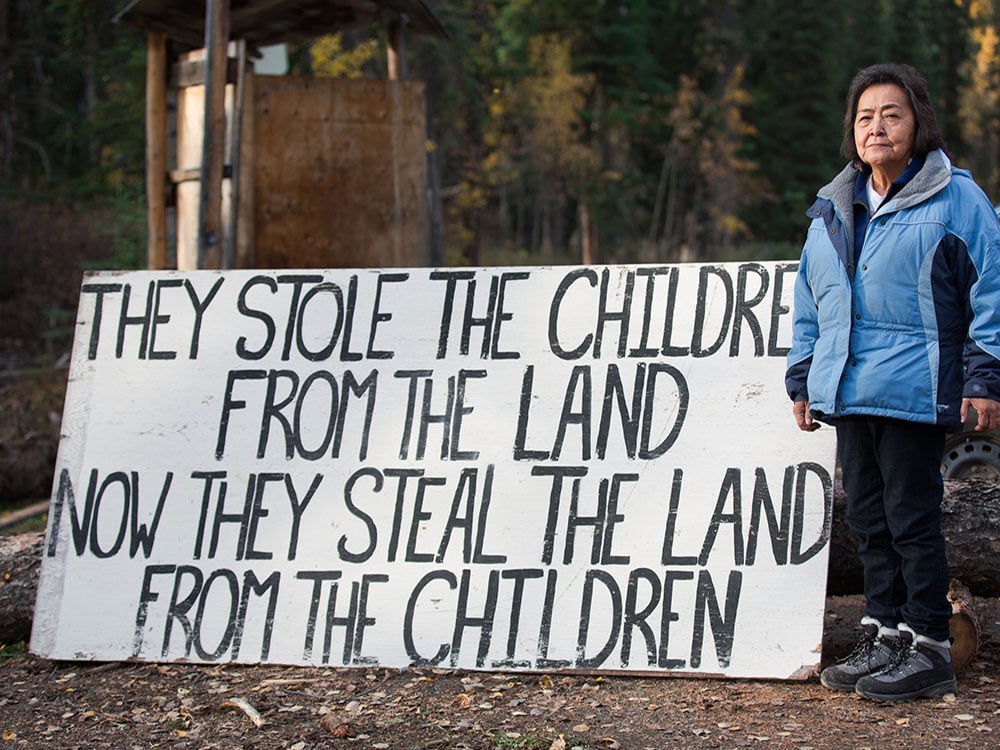 Elder Rita Louie is standing in a light blue winter jacket, black pants and hiking boots to the right of a large sign at a blockade. The sign reads “They stole the children from the land. Now they steal the land from the children.”
