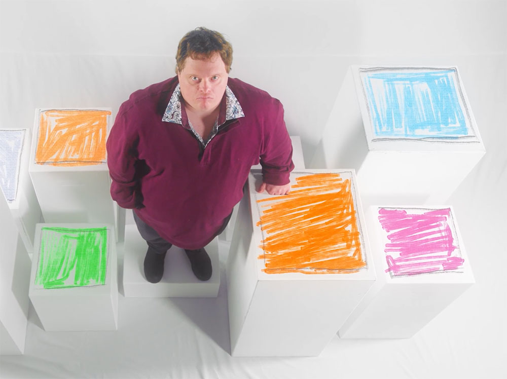 Niall McNeil is wearing a burgundy pullover and looking up at the camera. He is standing on a set featuring white boxes with colourful tops hand-coloured in marker.
