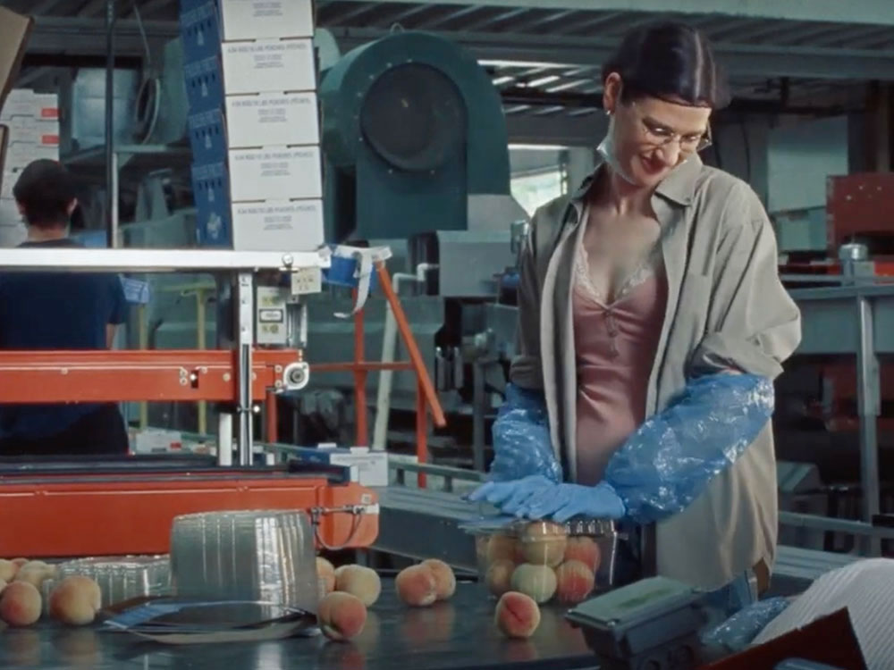 A woman stands inside a peach processing plant. She is wearing an oversized beige workshirt over a peach camisole. Her brown hair is under a hairnet. She is looking down.