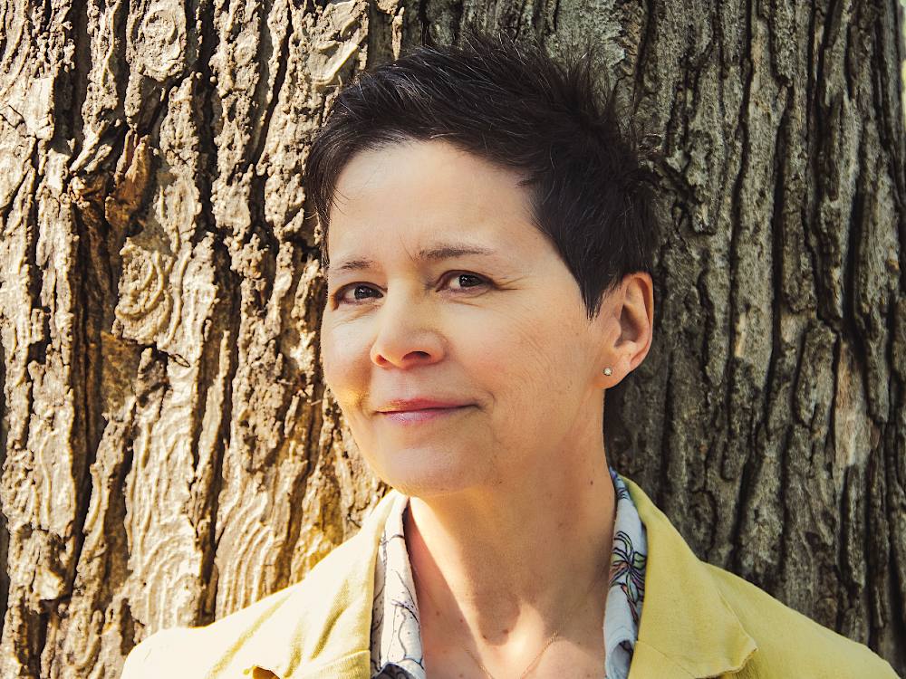 Ann-Marie MacDonald has short dark hair. She is standing against the trunk of a tree, looking at the camera with a peaceful smile. She is wearing a yellow collared jacket over a white shirt with a floral pattern.  