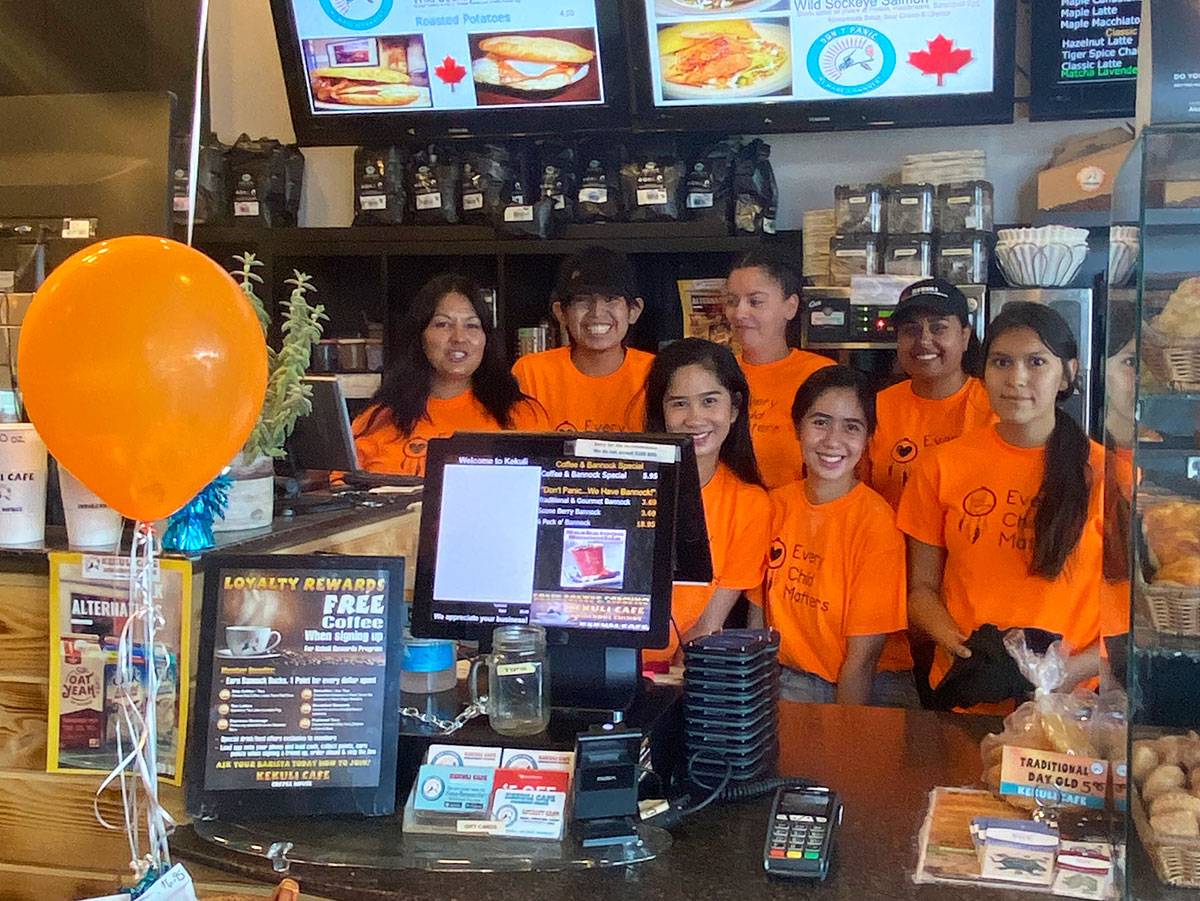 Seven employees stand behind a counter, flanked by display cases of bannock. They are all wearing orange shirts that read, “Every Child Matters.” 