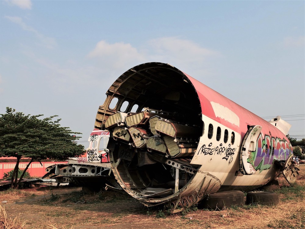 A decommissioned passenger plane stands in a dry grass field part of an ‘airplane graveyard’ in Bangkok, Thailand. Here, we see the deconstructed middle section of a red and white plane. Graffiti is on its right side and the interior is jammed with long pieces of scrap. In the background, the sky is blue. 