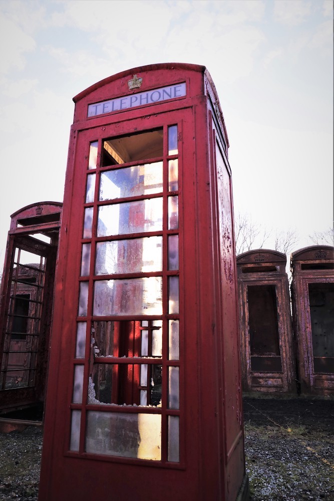 A red British telephone booth has broken windows up and down its side. It is standing in an outdoor lot amongst other old rusted red phone booths.