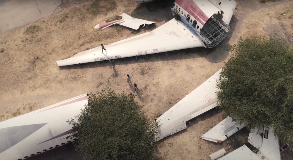 A bird’s eye view of a decommissioned passenger jet laid out across a field of burnt grass. The plane is white with red and blue markings. It’s in pieces across the field. Children are running and climbing on one of its wings. 