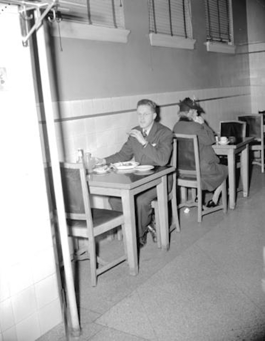 A black and white photo of a man in a suit sat at a two-person table eating what appears to be a sandwich.