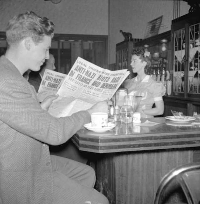A young man wearing a tweed suit sits at a lunch counter reading the paper. The headline is &ldquo;Anti-Nazi Rage in France and Denmark.&rdquo;