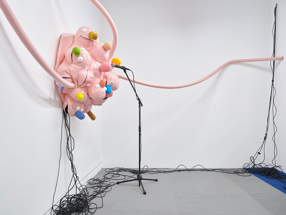 To the left of the frame, a baby-pink assembly of what appears to be children’s puppet heads with colourful soft fabric noses are arranged in a round cluster near a microphone. Black wires dangle from the puppet heads down a white wall onto a gray floor. Long baby-pink coils of fabric are in the foreground.