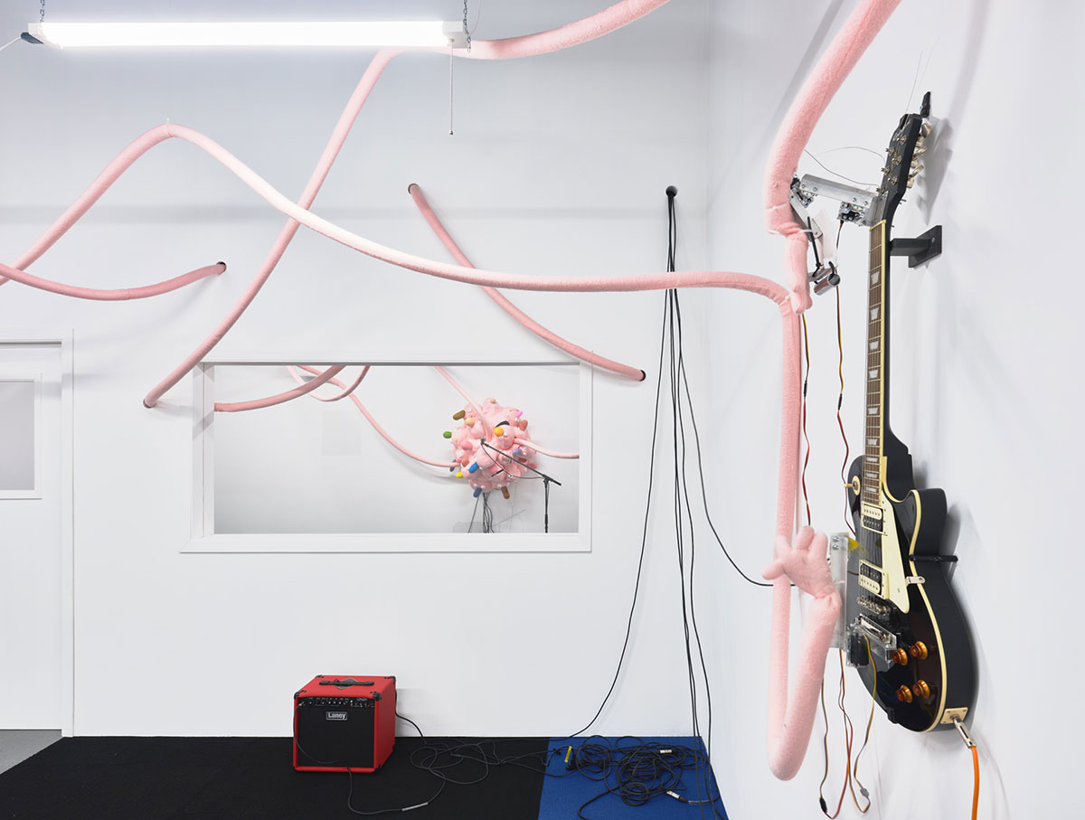 A room with white walls features baby-pink cylindrical tubes running across the ceiling and against the walls. To the right is an electric guitar. Through a window to another room is a faraway round cluster of puppets facing a microphone. Their features are indistinguishable from this distance.