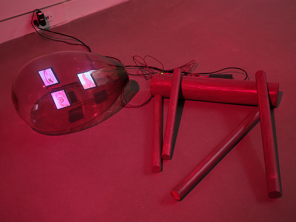 A red robot is collapsed on its side on the floor, dimly lit in red. The robot’s body, appendages facing down to the right of the screen, is comprised of cylindrical tubes. To the left, its glass lightbulb-shaped head contains digital images of two eyes and a mouth. 