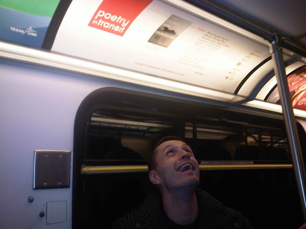 Rob Taylor sits on a bus, happily looking upwards. His Poetry in Transit placard is located in the ad area above his head.