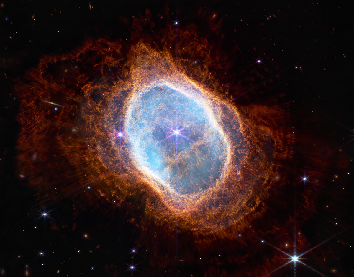 A bright white star at the centre of a nebula radiates out to light blue circular shape with an outer white ring that gives way to reddish exterior that fades into a black galactic sky.