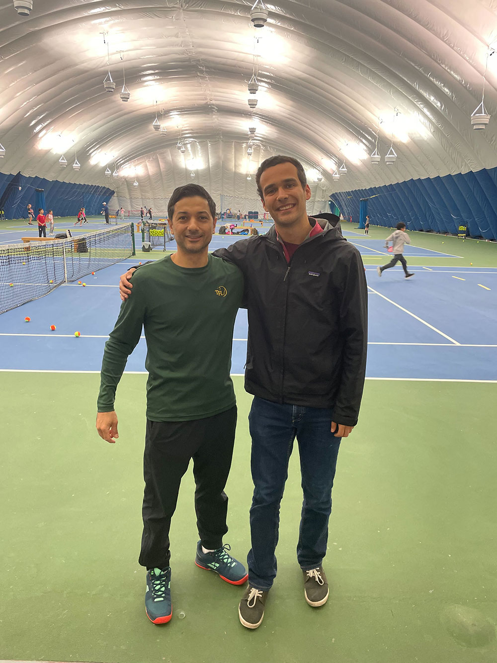Hassan Askari, left, and Josh Kozelj, right stand with their arms around each other’s shoulders. Hassan is wearing a dark green long-sleeved t-shirt. Kozelj is wearing a black rain jacket. They are both standing on green turf at the Coquitlam Tennis Centre, an indoor tennis centre with a domed white ceiling visible above them. Behind them, kids practice tennis on blue indoor courts with white lines.