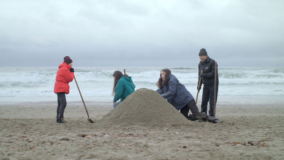 Four people are wearing winter attire on a beach. Whitecapped waves roll behind them. The people are creating a mound of sand on the beach. A woman on the left is leaning against a shovel in a bright red winter jacket and toque. Two younger women with long hair in teal and blue jackets work on the mound. An older man stands to the right, holding a shovel wearing a black jacket, pants and a toque.