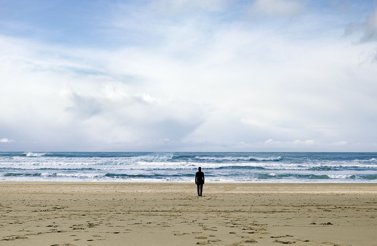A black silhouette stands on a sandy beach against a bright blue horizon with whitecapped waves rolling towards the beach. The sky is blue and full of white fluffy clouds.