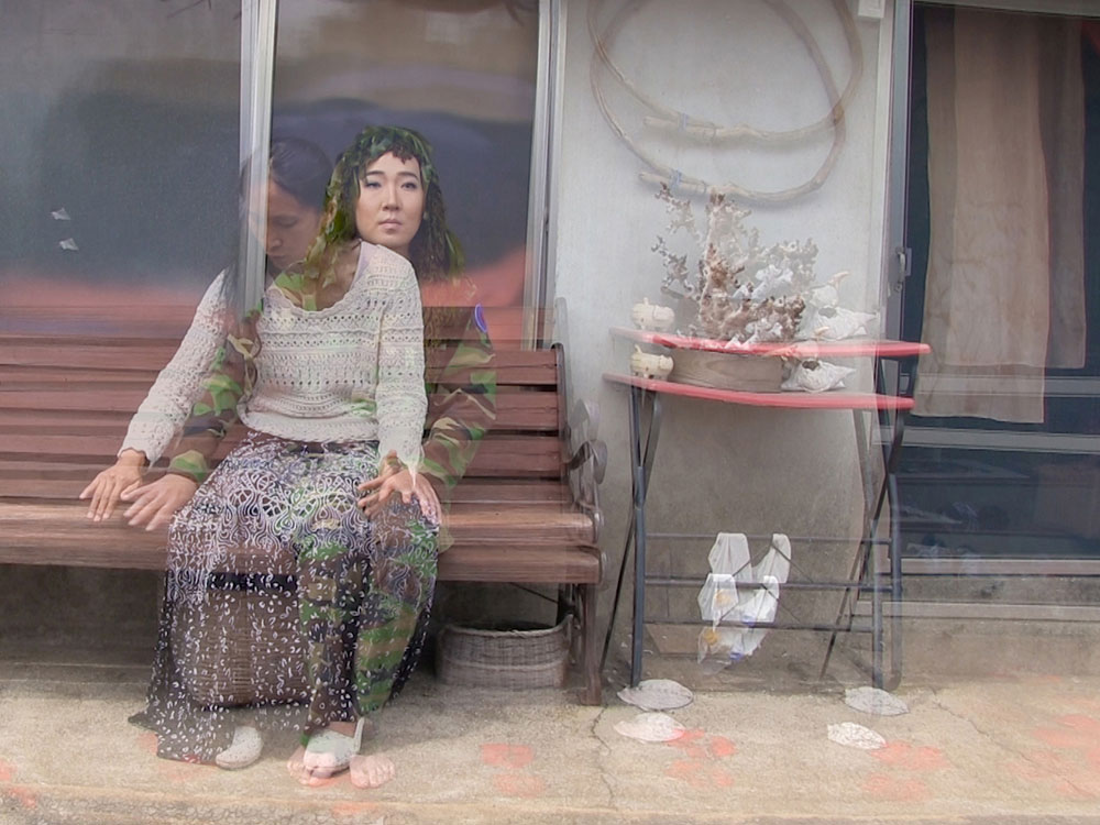 Two overlapping video images of women in different outfits occupy a brown bench in front of a screen door. An image of a woman seated in green military fatigues is layered under an image of a woman seated and looking down in a white sweater and long navy skirt. To their right is a double image of a red side table and a curtained door.