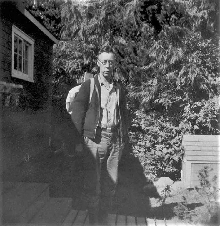 A black-and-white photo of a man standing in front of a shack, wearing circular glasses.