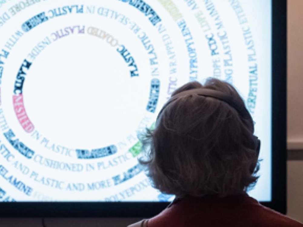 A woman with short grey hair is wearing headphones and looking at a large white screen with a spiral digital image in which the word “plastic” is spelled in capital letters. We can see the back of the woman’s head. She is standing in a dim space at the Surrey Art Gallery.