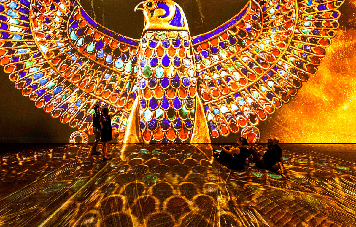 A gold floor foregrounds a colouful projection of a falcon in the style of ancient Eygyptian artwork. The falcon is coloured with blue, red, green, white and black. A couple stands to the left of the falcon, with one person pointing to the artwork. Another couple is sitting casually on the floor.