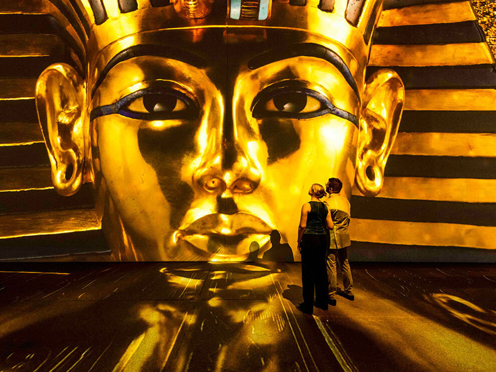 A couple stands to the right of a large-scale art installation featuring King Tutankhamun's famous gold death mask. The mask is a gold floor-to-ceiling projection of the ancient Egyptian king's face in gold, his eyes lined in black kohl.