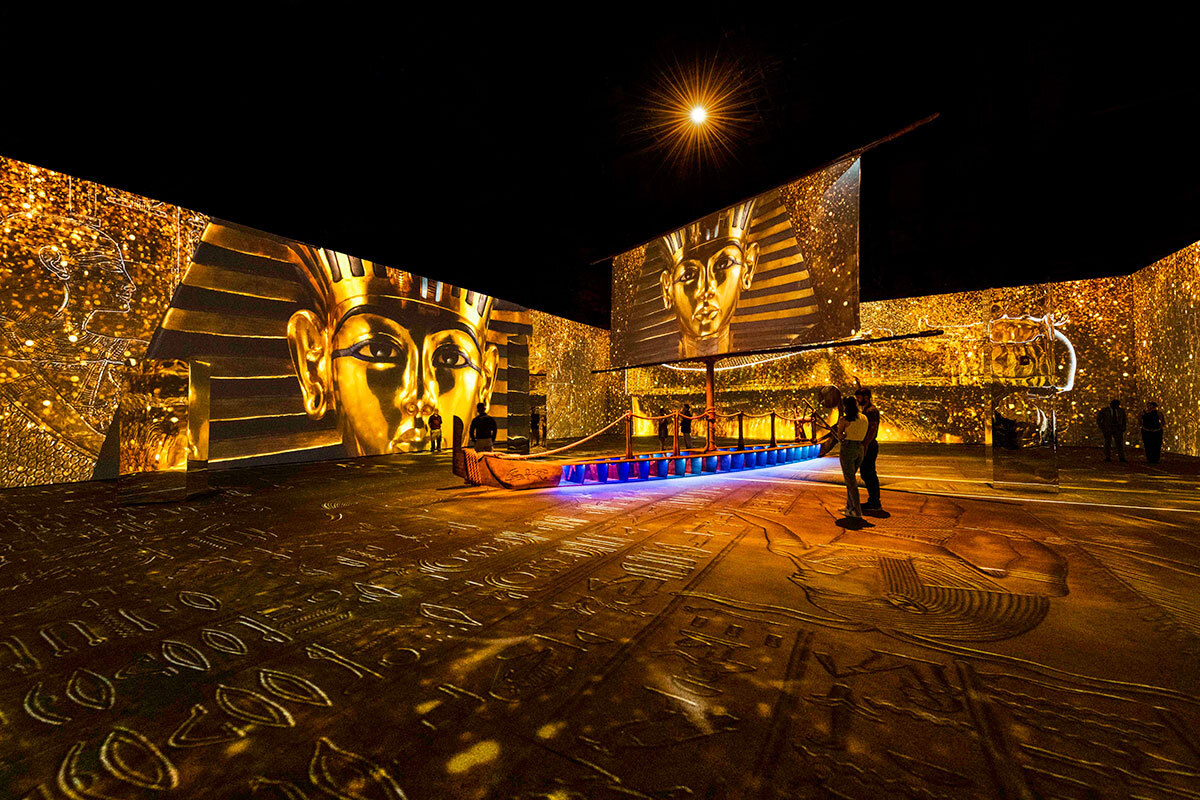A large room features a gold floor cast in hieroglyphics. King Tutankhamun’s famous gold death mask is projected on two screens in the room. In the background are projected screens of Egyptian artwork with an overlay of gold sparkles. Silhouettes indicate a small group of people scattered throughout the art exhibition.