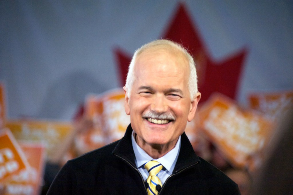 Jack Layton smiles towards the camera. People holding orange NDP signs are blurrily visible behind him.