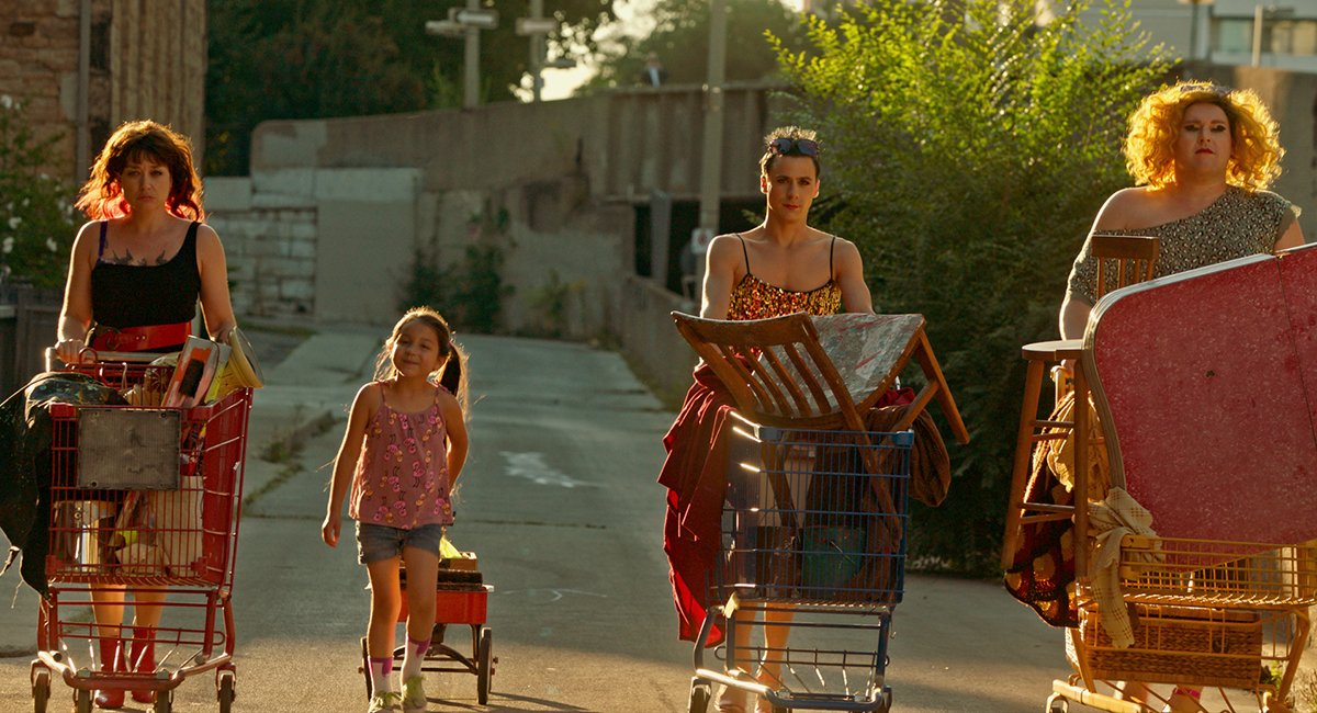 Three adults are pushing shopping carts full of furniture down an alley in Montreal at Sunset. Keris Hope Hill, who plays six-year-old Rosie in the film, is wearing pigtails and pulling a red wagon behind her.