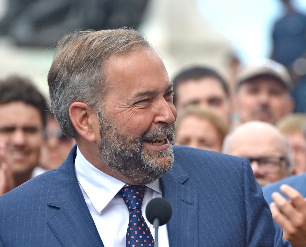 Tom Mulcair laughs with someone off-camera to his left. People are blurrily visible behind him.
