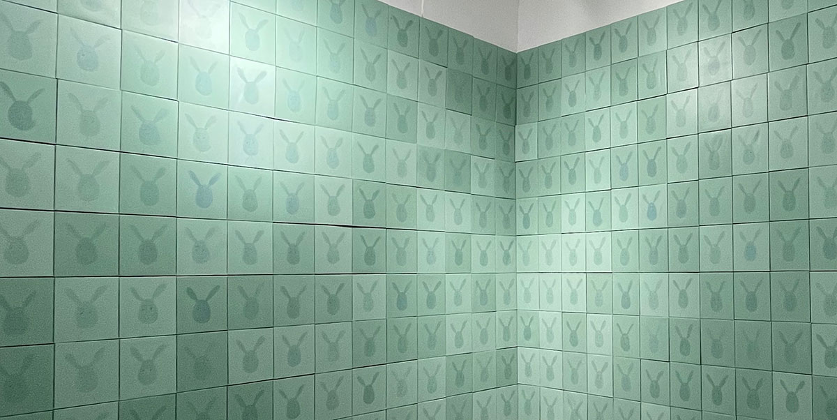 A close-up of Bill Pechet’s Who’s There features a corner of a tiled wall featuring many seemingly-identical seafoam tiles with a dark illustrated silhouette of a bunny’s head. Look closer, and each rabbit’s face is hand-painted with a distinctive facial expression.