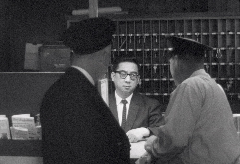 A black and white photo from 1964 shows a Chinese man wearing glasses and a suit and tie. He is standing behind a desk. There is a wall of hotel keys hanging behind him. In the foreground are two police officers in uniform with their backs turned to the camera. They are talking to the Chinese man.