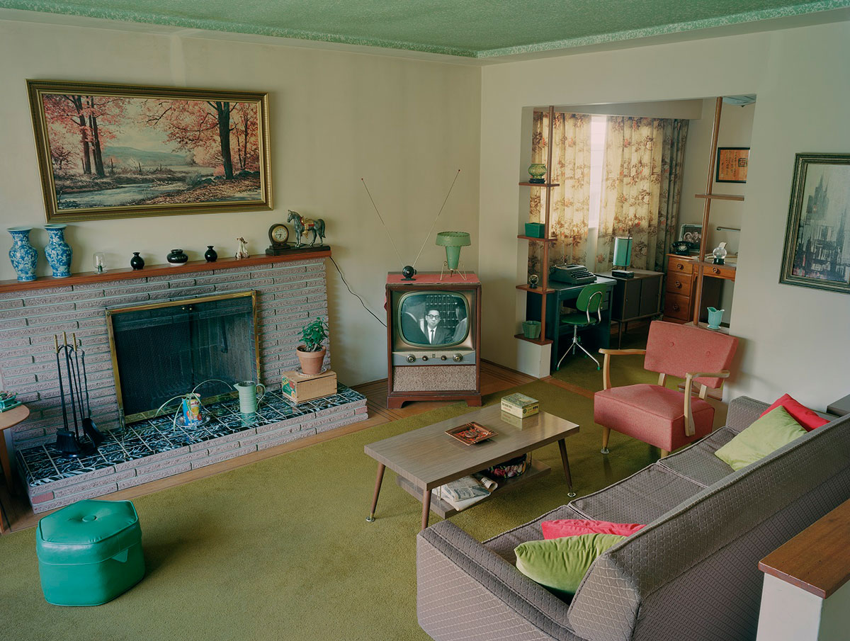 The living room of a classic Vancouver house called the mid-century builder. The TV, showing black-and-white news footage of a Chinese man in a suit and tie, is in the centre of the room. On the left of it is a fireplace. On the right of the TV is a glimpse into a study with a typewriter. This recreation is set in 1964, which we can tell from the mid-century furniture and green-tinted carpet and ceiling. This looks like an average working-class home in Canada, but there are hints like the porcelain vases and Chinese calligraphy that say this is a Chinese-Canadian household.
