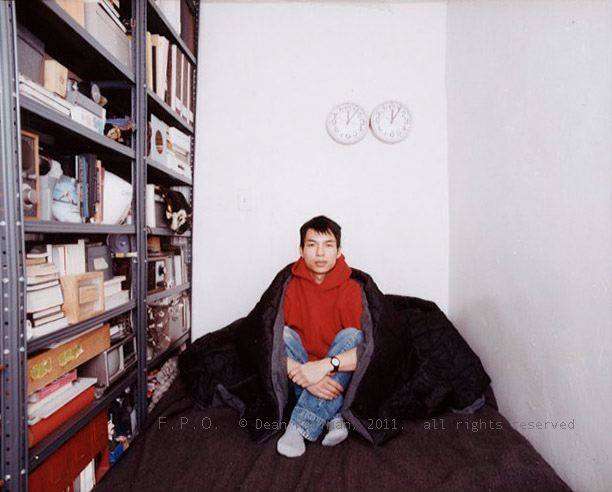 Tobias Wong sitting on a bed in a red hoodie, blue jeans and grey socks with a black Kevlar duvet around his shoulders. The bed is against a white wall to the right of the frame and a crowded bookshelf to the left of the frame. Above the bed are two white analogue plastic clocks.