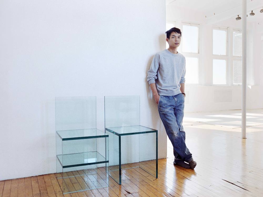 Tobias Wong is standing against the corner of a white wall. He is wearing a light grey crewneck sweatshirt over a white T-shirt and jeans. He is looking at the camera with a neutral expression. To his left are two glass chairs modelled after artist Donald Judd’s Chairs 84, which were made from flat pine boards. Wong’s chairs are made of panes of clear turquoise glass.