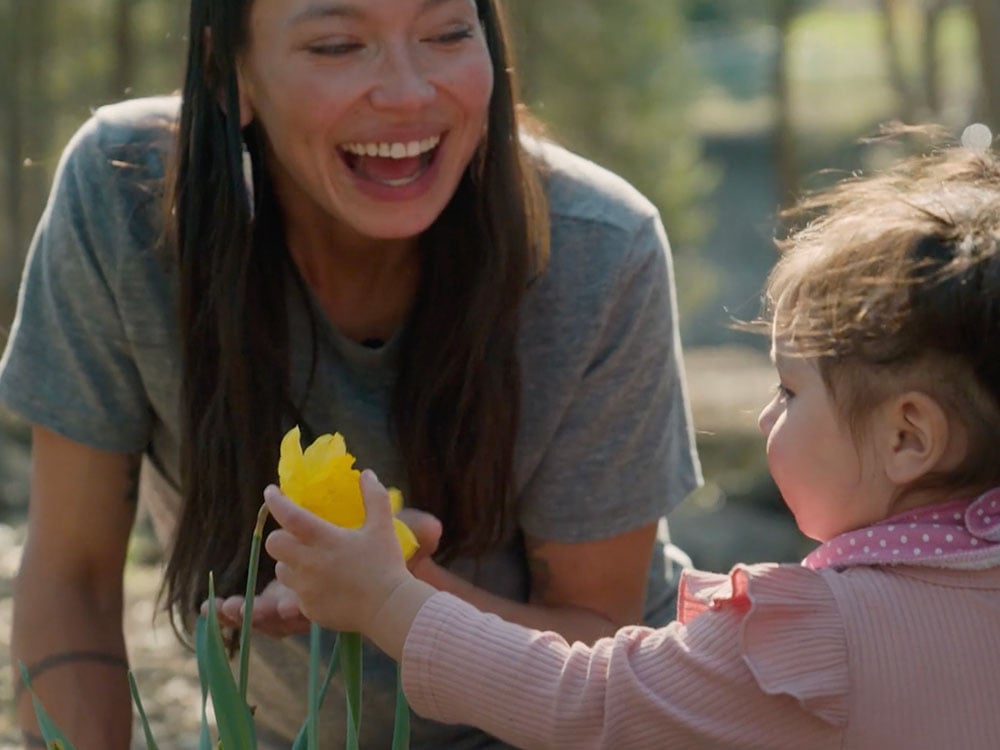 An Anishinaabe woman with long dark hair and wearing a grey T-shirt is smiling widely at her toddler daughter dressed in pink to the right of the frame. Her daughter is holding the yellow blossom of a daffodil in her hands. The two are outdoors in a sunny wooded area.
