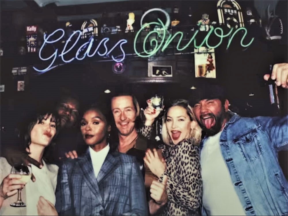 A polaroid party photo depicts a group of five friends, holding drinks and mugging for the camera. The words “Glass onion” hang across the top of the frame as a blue and green neon sign.