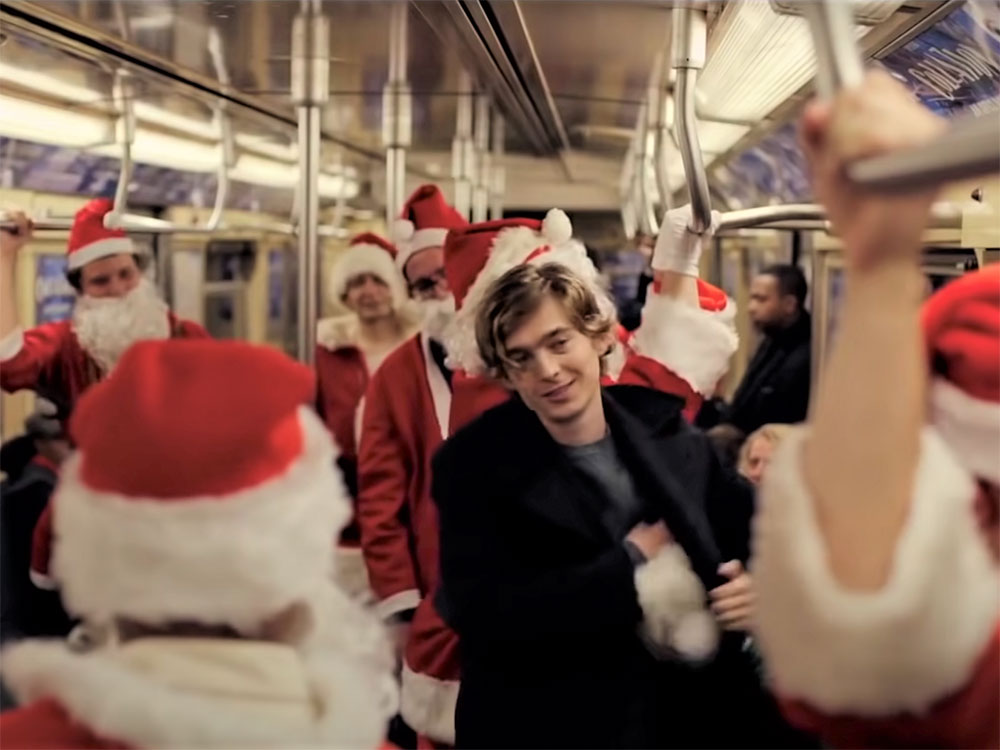 A young man with shaggy blonde hair and a black jacket stands on a crowded subway surrounded by people in Santa suits.
