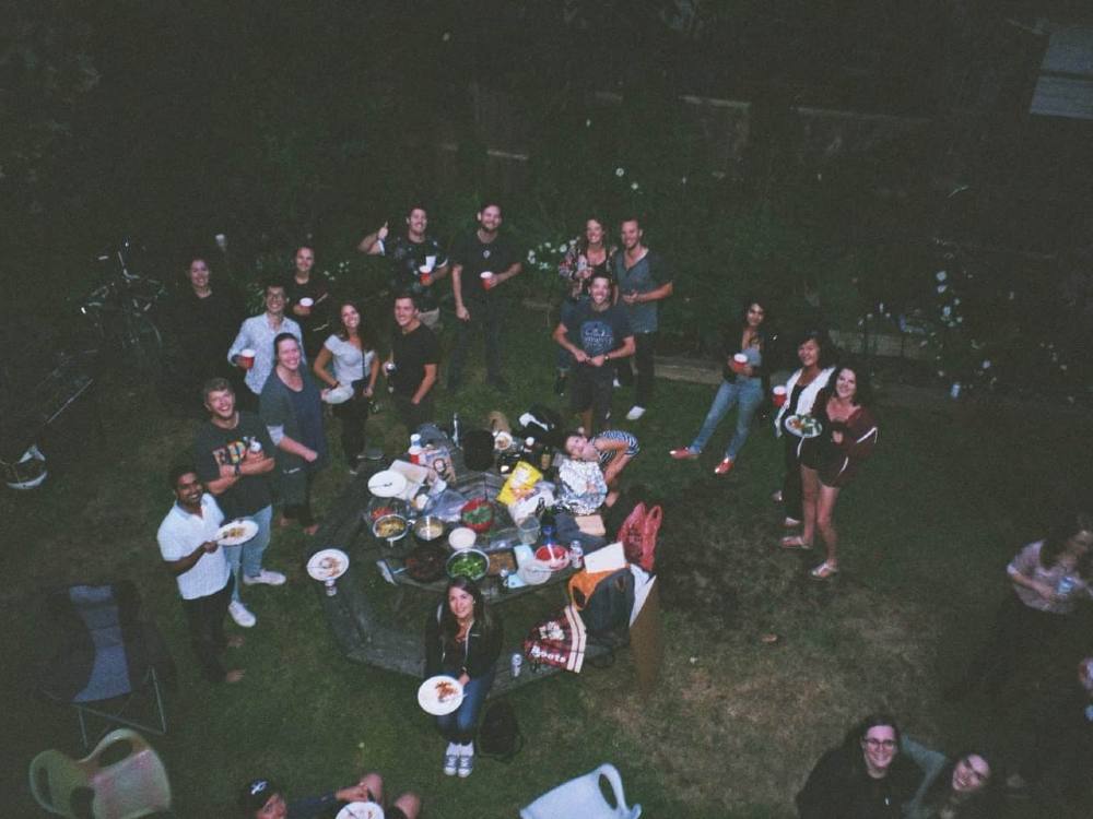 An aerial shot of about two dozen people standing around with plates and drinks in a backyard. Most people are smiling up at the camera. It’s dusk, and the light is low.