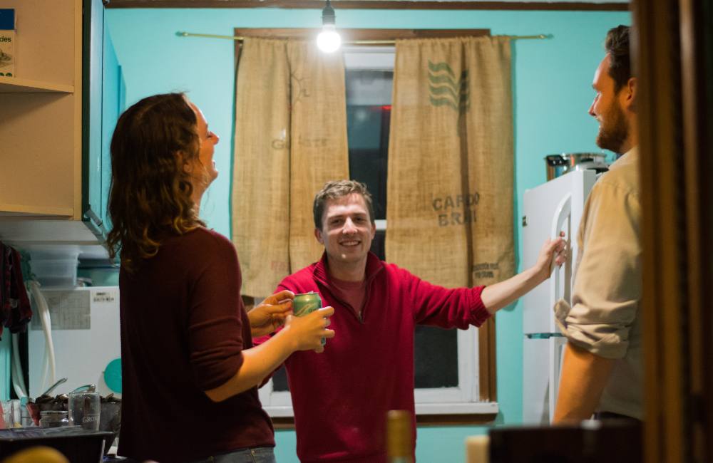 Three people stand in a turquoise-painted kitchen. On the left, Michelle is laughing and looking at Tom, in the centre, who is looking at the camera.