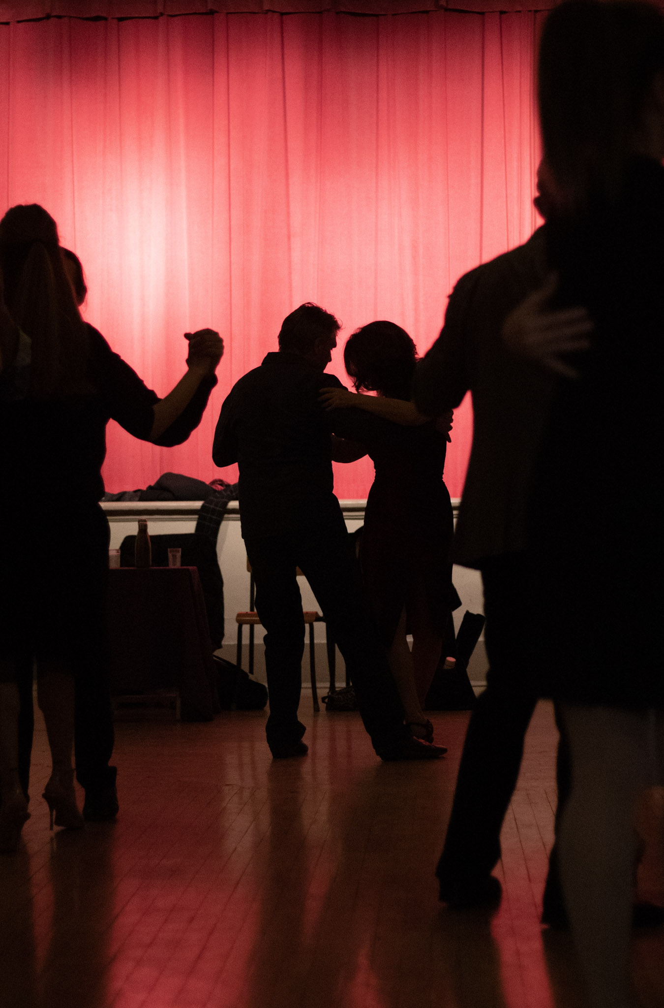  A couple dancing are black silhouettes against the deep red curtain. They are framed by other dancing silhouettes and mirrored by their reflection in the polished floor. 