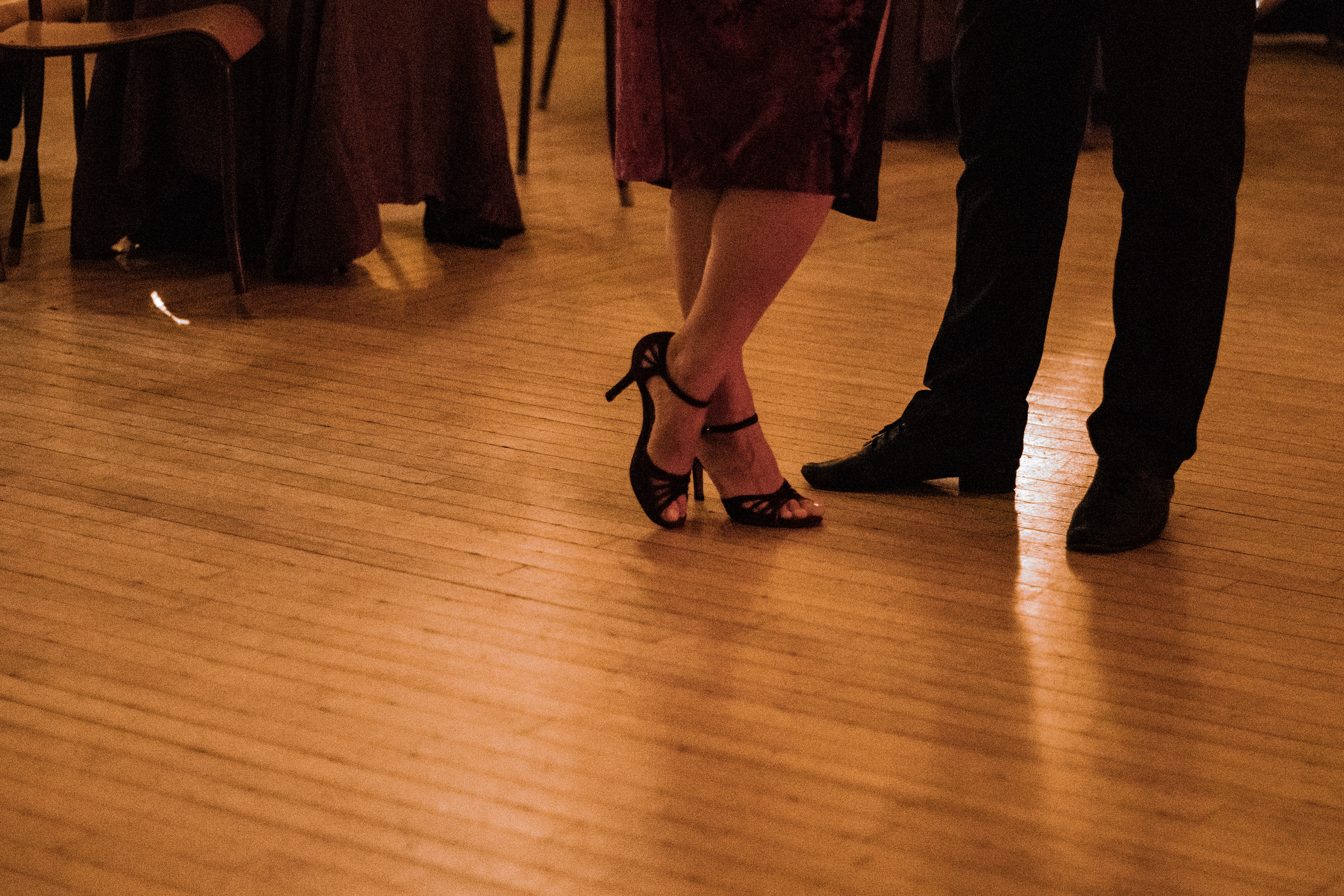 A woman and man’s feet rest upon hardwood floors that shine with a soft yellow light. The woman’s feet, wearing red high-heels, are crossed over each other, the toe of the crossed left foot rests on the ground. The heel is pointed in the air. 