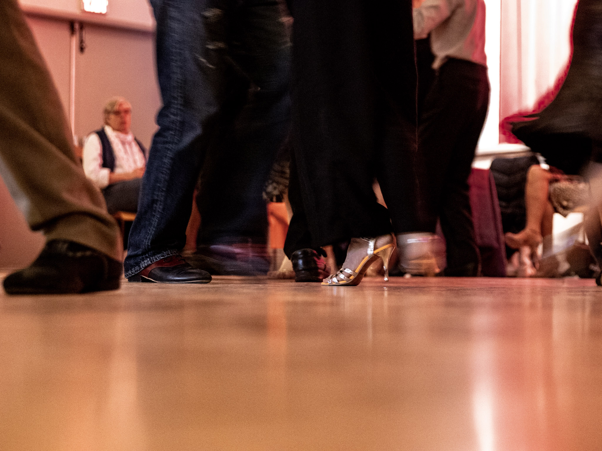 A photograph capturing the movement of those on the dance floor. Two feet are motionless while the out-of-focus whirl of moving legs, feet and bodies surrounds them. 
