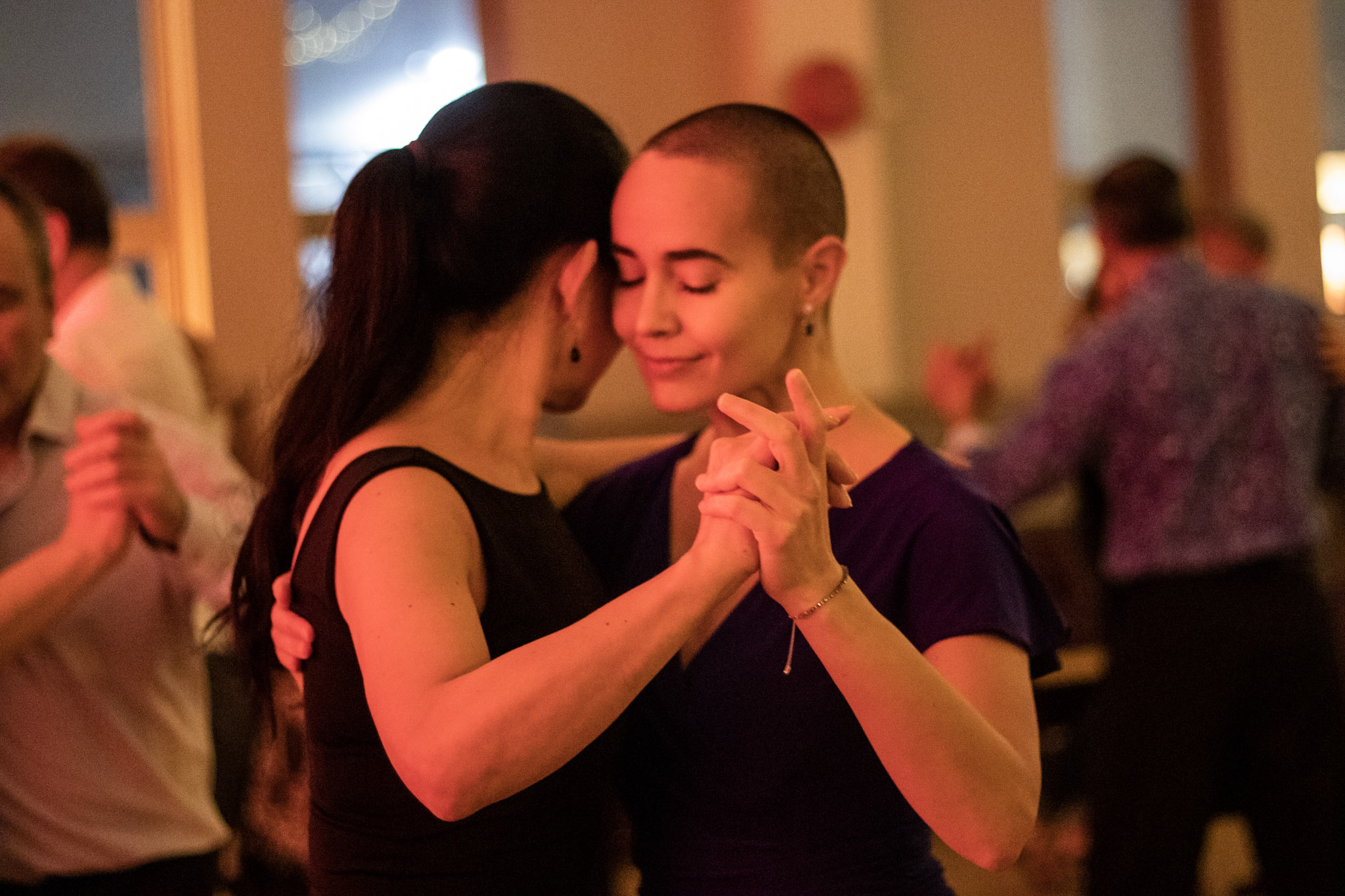 Two women dance together, cheek to cheek. The camera focus’ on the hands clasped together. In the background, slightly out of focus is the face of the woman who leads, her eyes closed. She is smiling. 