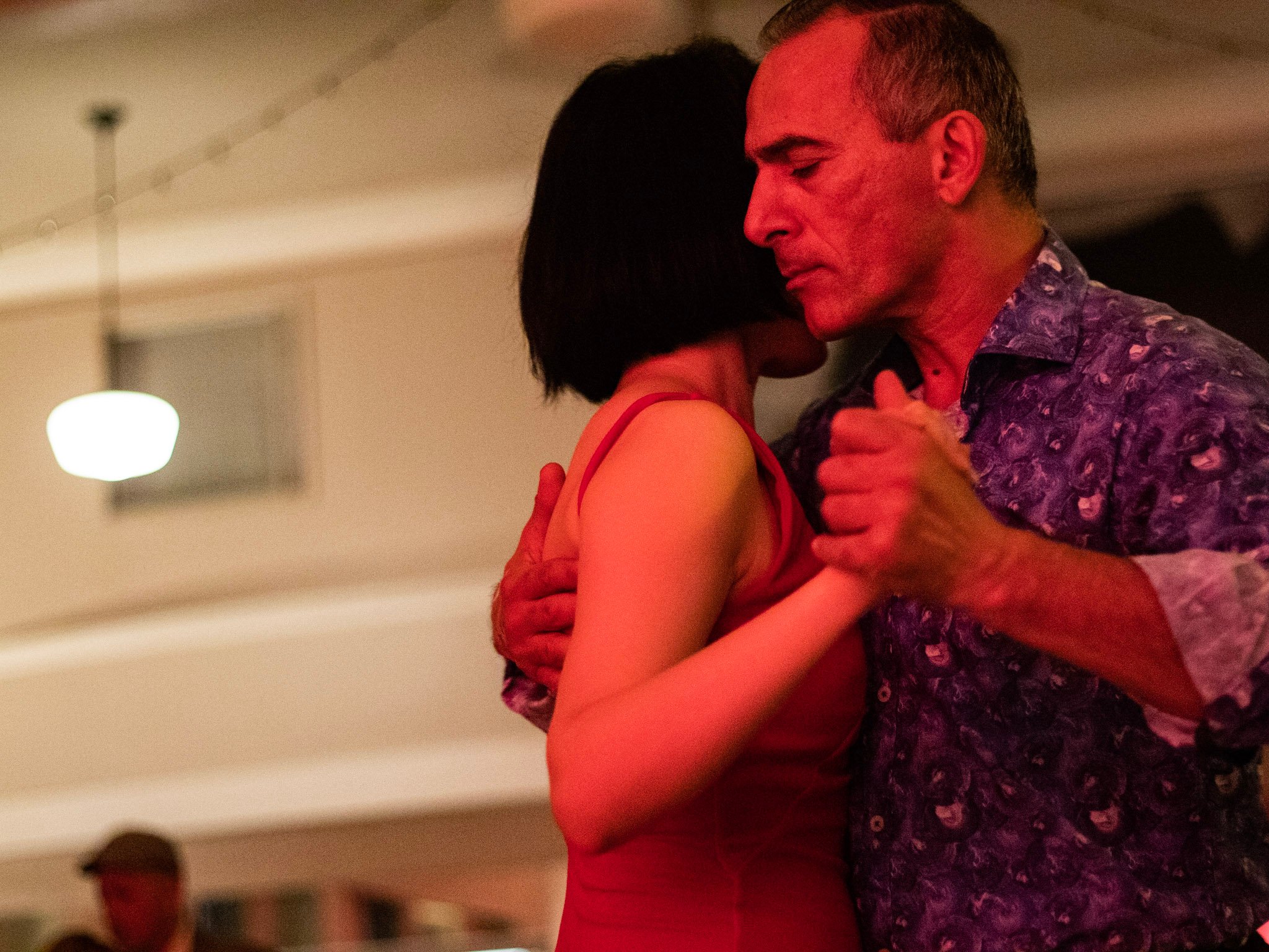 ID: A close-up photograph of a man and a woman dancing. The woman’s face is hidden behind the man’s face, his eyes are closed as their chests are pressed together and their hands clasped.