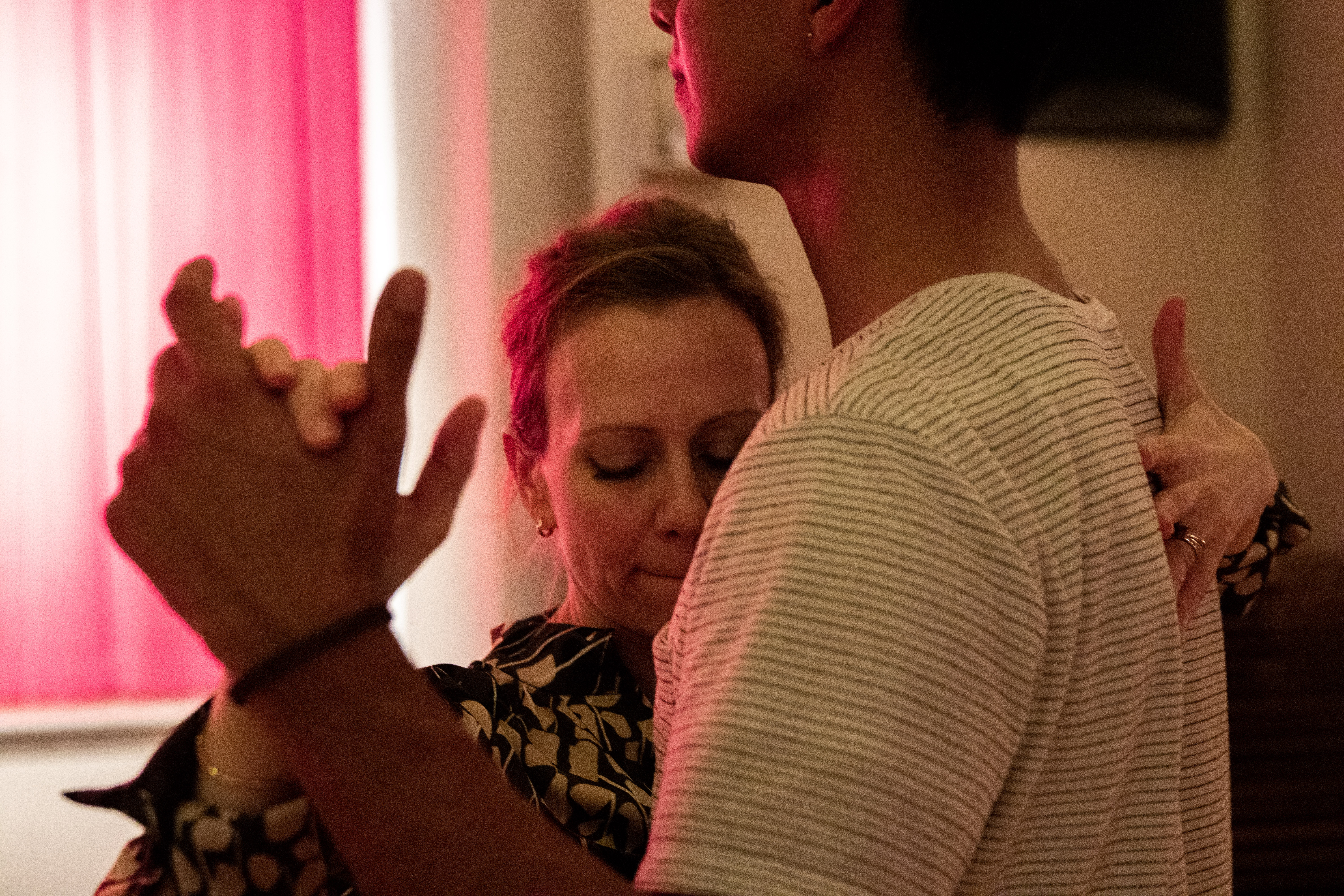 A close-up of a woman and young man dancing. His face is cut off by the frame, but her face presses into his chest. Her eyes are closed, her mouth pressed in concentration. The red light from a curtain graces the left side of her face. 
