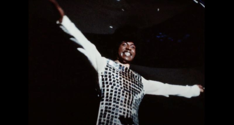 A Black man in a white long-sleeved top with shiny silver tiles spreads his arms and smiles at an audience out of frame. The background is dark.