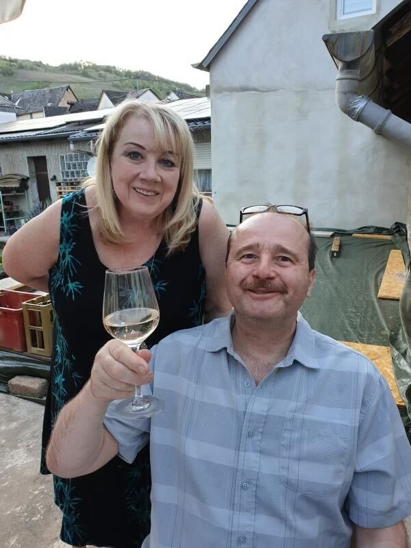 Paula Dauns has blonde hair, wearing a dark sleeveless dress and is standing towards the left of the frame over her husband Ralf’s right shoulder. Ralf is wearing a light blue shirt and raising a glass of white wine.