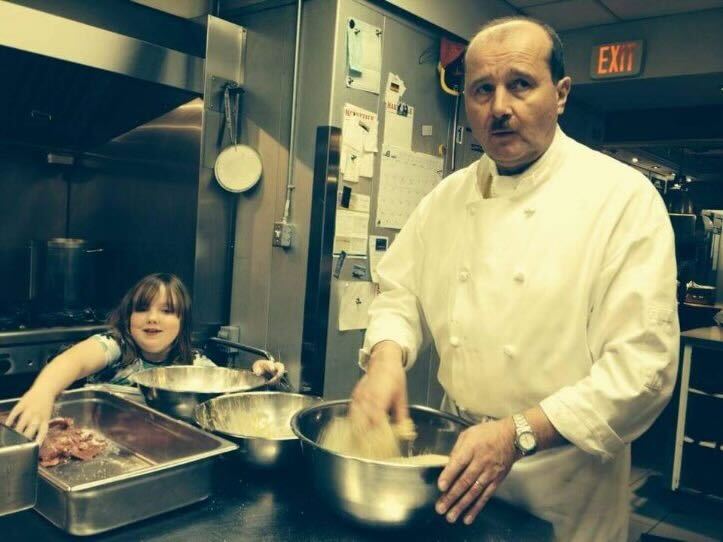 A young girl stands with Ralf Dauns at a stainless steel counter in the Soup Meister kitchen, where they both have their hands in a floury mixture in stainless steel bowls. 