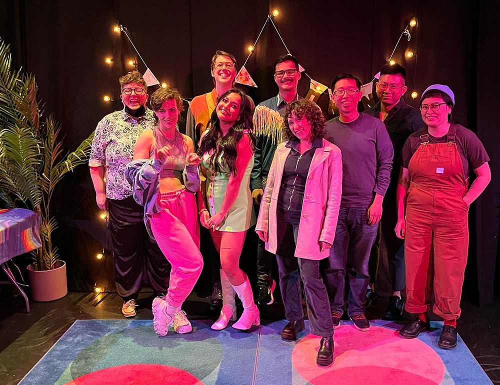 A group of comedians stands together for a group photo against a black wall with fairy lights and bunting. They are smiling at the camera.